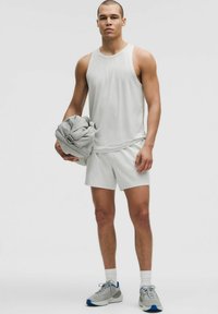 Top deportivo sin mangas de color gris claro y pantalones cortos, con textura suave. Modelo sosteniendo una bolsa de gimnasio gris. Zapatillas deportivas con acentos azules y calcetines blancos.