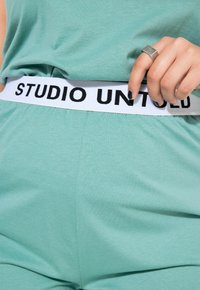 Ensemble de détente vert menthe avec une taille élastique blanche portant l'inscription "STUDIO UNTOLD" en noir. Texture lisse et coupe décontractée.
