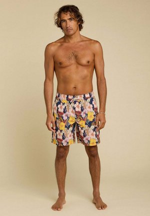 Homme aux cheveux bouclés mi-longs, debout pieds nus, portant un short de bain à motifs floraux aux teintes jaunes, roses et bleues, sur un fond uni.