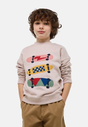 Enfant aux cheveux bouclés portant un sweat-shirt beige imprimé de trois planches à roulettes colorées et un pantalon marron, les mains dans les poches, fond neutre.
