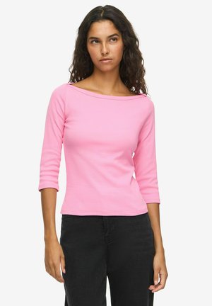 Woman with dark curly hair wearing a pink boat neck top with three-quarter sleeves and black pants, standing against a plain white background.