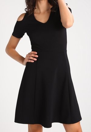 Woman wearing a black knee-length dress with cold shoulder cutouts and a V-neckline, standing with one hand on her hip.