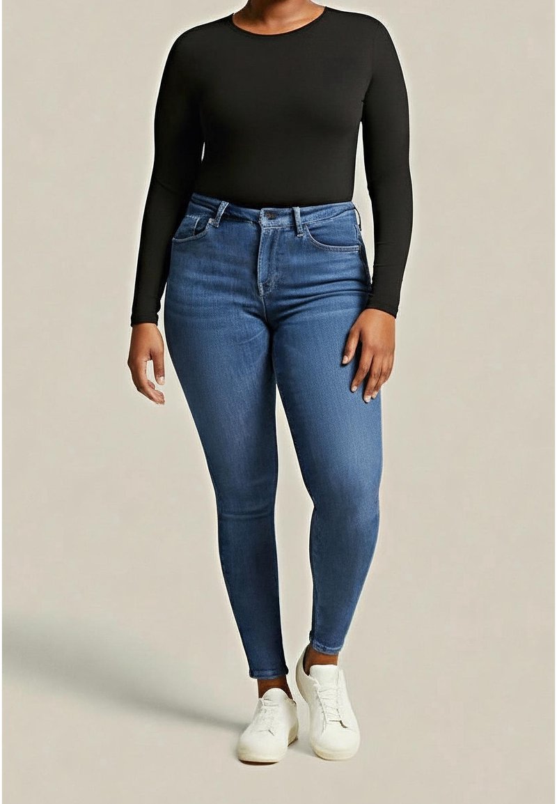 Personne portant un haut noir ajusté à manches longues, un jean skinny bleu taille haute et des baskets blanches, debout sur un fond neutre.