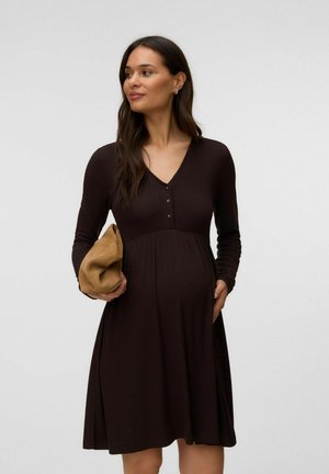 Gravid kvinde iført en mørkebrun kjole med lange ærmer, holder en lys tan clutch, har den ene hånd på maven og kigger til siden.