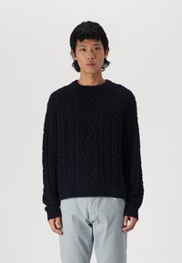 Dunkelblauer Kabelstrickpullover mit Rundhalsausschnitt und gerippten Bündchen, der strukturierte Muster aufweist, die vertikal über den Stoff verlaufen.