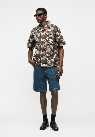 Άνδρας που φοράει καφέ και μπεζ κοντομάνικο πουκάμισο παραλλαγής, μπλε τζιν σορτς μέχρι το γόνατο, μαύρες κάλτσες, μαύρα παπούτσια και γυαλιά ηλίου, στέκεται με το ένα χέρι στην τσέπη.