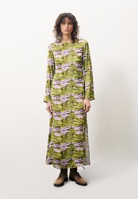 Maxikleid aus grünem Stoff mit einem Muster aus Bergen und Bäumen in Pink. Es hat lange Ärmel und eine gerade Silhouette, kombiniert mit braunen Schnürstiefeln.