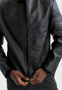 Veste en cuir noire avec détail matelassé aux épaules, fermeture éclair avant, deux poches latérales et une texture lisse. Portée sur un haut en maille noir.