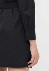 Abito nero con cintura, realizzato in un tessuto morbido e leggero; presenta un polsino con bottoni e uno spacco all'orlo, mostrando un design semplice e pulito.