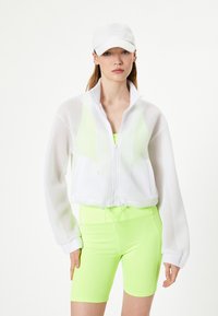 Hvid mesh zip-jakke over en neon grøn tætsiddende top og matchende shorts. Funktioner inkluderer en høj krave og snoretræk i taljen. Accessories omfatter en hvid kasket.