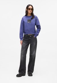 Purple cropped sweatshirt with a black patch logo, paired with high-waisted black jeans and chunky black shoes. Model wears dark sunglasses.