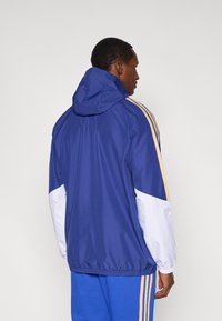 Modrá športová bunda s kapucňou, so bielymi a zlatými akcentmi na rukávoch. Vyrobená z ľahkého materiálu s elastickými manžetami a lemom.