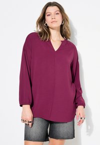 Pflaumenfarbene Bluse mit langen Ärmeln, V-Ausschnitt und gerafften Schultern, kombiniert mit grauen Jeansshorts mit fransigem Saum.