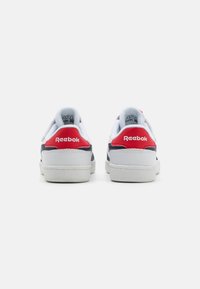 Zapatillas blancas con un empeine de cuero suave, detalles en azul marino y pestañas rojas en el talón. Cuenta con una suela de goma texturizada y un detalle de logo en la parte posterior.
