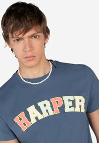 Camiseta de algodón azul con "HARPER" en letras en negrita de color amarillo, rojo y rosa. La lleva puesta con un collar de cuentas blancas.