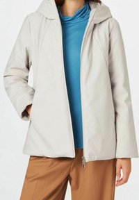 Veste gris clair à fermeture éclair avec capuche, présentant une texture lisse et des manches raglan, superposée sur un col roulé bleu sarcelle. Pantalon marron en dessous.