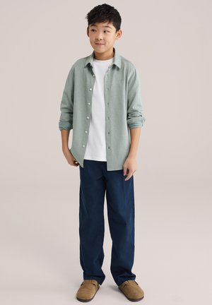 Svetlozelená košeľa na zapínanie cez bielu tričko, doplnená o tmavomodré nohavice a béžové slip-on topánky. Oblečenie má uvoľnené siluety.