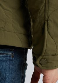 Olivgrüne gesteppte Jacke mit schmalem Bündchen, schwarzem Knopfakzent und strukturiertem Stoff, kombiniert mit blauen Jeans.