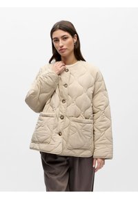 Γυναίκα με μπεζ καπιτονέ σακάκι με κουμπιά και μεγάλες μπροστινές τσέπες, φορεί σκούρο φαρδύ παντελόνι και ποζάρει μπροστά σε λευκό φόντο.