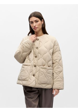 Vrouw draagt beige gewatteerd jack met knopen en grote voorzakken en donkere losse broek, poseert tegen een witte achtergrond.