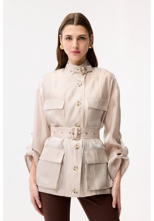 Kvinde iført beige jakke med bælte og store lommer samt guldfarvede øreringe, stående foran en ensfarvet hvid baggrund.