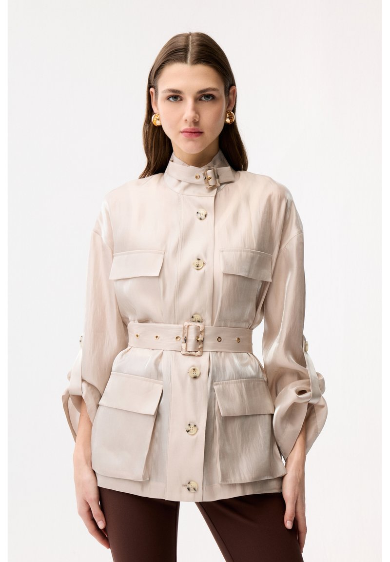 Woman wearing beige belted jacket with large pockets and gold hoop earrings, standing against a plain white background.