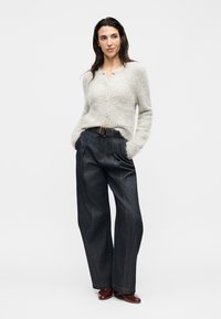 Femme portant un cardigan blanc duveteux boutonné, un jean sombre à jambes larges avec une ceinture et des chaussures marron à talons, debout avec les mains dans les poches.