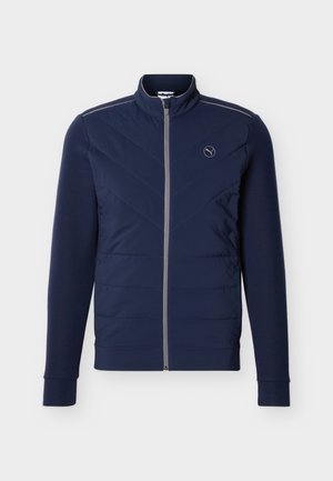 Tumši zila josta virsū ar chevron šūšanu, augstu apkakli, garām piedurknēm un mazu Puma logo kreisajā krūtīs.