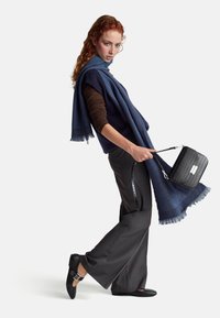 Czarna torebka o teksturze krokodyla z srebrnymi elementami. Modelka nosi ciemnoniebieski szalik i brązową bluzkę, zestawione z szarymi spodniami i czarnymi butami.