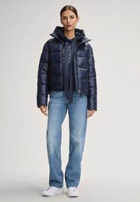 Giacca imbottita blu navy con chiusura a zip e cappuccio. Indossata sopra un felpa blu navy e pantaloni jeans larghi azzurri. Sneakers bianche.