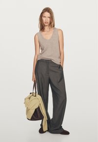 Žena v béžovém bezrukávovém topu a tmavě šedých širokých kalhotách, držící tmavohnědou tašku s světle žlutým svetrem přehozeným přes ni.