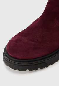 Burgundowe zamszowe botki za kostkę z okrągłym czubkiem i grubą czarną gumową podeszwą o teksturowanej powierzchni i wyraźnym wzorze bieżnika.