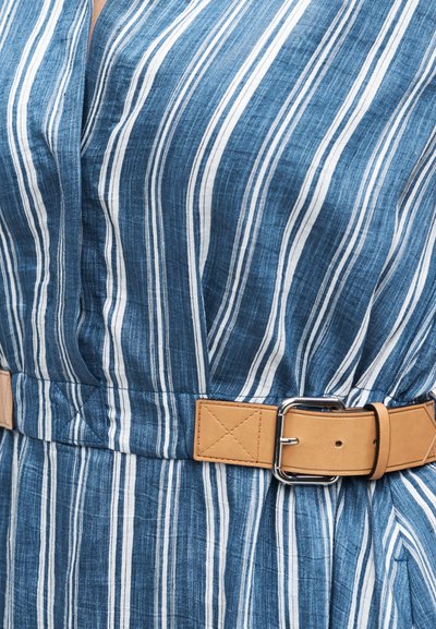 Γυναίκα που φοράει μπλε και λευκό φόρεμα με κάθετες ρίγες και ταμπά δερμάτινη ζώνη στερεωμένη στη μέση.
