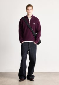 Μπορντό πουλόβερ με ψηλό γιακά και φερμουάρ, συνδυασμένο με σκούρο μπλε τζιν παντελόνι ευρείας γραμμής. Μαύρη τσάντα χιαστί, καφέ παπούτσια. Απλός, υφασμάτινος σχεδιασμός.