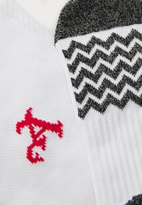 Weiße Socken mit einem roten Logo und einem gemusterten schwarzen Bündchen, das Zickzacklinien aufweist. Die Textur ist glatt, mit einem Rippenteil am Knöchel.