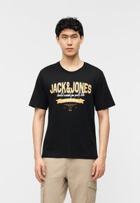 Νεαρός άνδρας με κοντά σκούρα μαλλιά που φοράει μαύρο T-shirt Jack & Jones και μπεζ παντελόνι cargo, στέκεται μπροστά σε λευκό απλό φόντο.