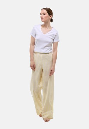 Chemise blanche à manches courtes en V associée à un pantalon large jaune clair avec de fines rayures blanches. Le modèle se tient pieds nus, les mains dans une position détendue.