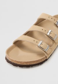 Beige sandal with three adjustable straps, silver buckles, and a brown textured sole. Made from synthetic material with a smooth upper.