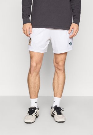 Hvite atletiske shorts med et teksturert mønster, en brodert logo, kombinert med grå sko og hvite sokker med en logo. En mørk, langermet topp er synlig.