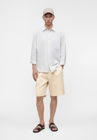 Homme portant une casquette beige, une chemise bleu clair à manches longues avec boutons, un short beige longueur genou et des sandales marron foncé, debout avec une main dans la poche.