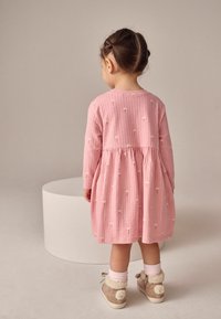 Růžové dlouhé šaty s texturovaným vzorem, zdobené malými žlutými květinovými akcenty a s nabráním v pase. Viditelné béžové boty s kožíškem.