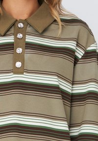 Polo rayé en vert olive avec des rayures blanches et brunes. Présente un col et cinq boutons décoratifs. Texture du tissu lisse.