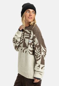 Jonge vrouw in een bruin en crèmekleurig patroon trui, zwarte beanie en donkere broek, die opzij kijkt met een lichte glimlach.