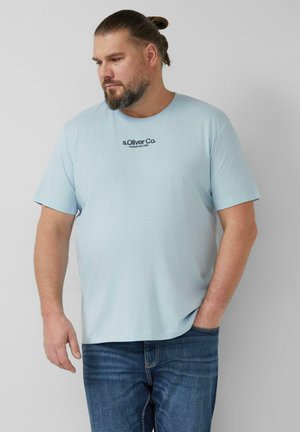 Homme barbu aux cheveux attachés, portant un t-shirt bleu clair de la marque s.Oliver Co. et un jean bleu, debout avec une main dans la poche.