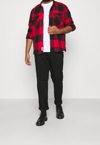 Camicia flanellata a quadri rossi e neri, pantaloni neri e scarpe nere. T-shirt a collo rotondo bianca sotto, con due tasche sul petto nella camicia.
