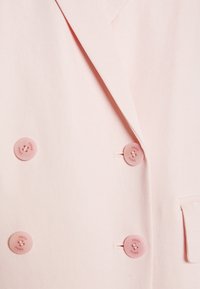 Světlé růžové sako s dvojitým zapínáním a velkými růžovými knoflíky, na přední straně s jednou kapsou. Tkanina má hladký, jemně strukturovaný povrch.