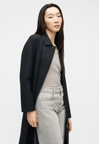 Νεαρή γυναίκα με μαύρο παλτό πάνω από μπεζ ριγωτό αμάνικο τοπ και ανοιχτό γκρι τζιν ψηλόμεσο, που στέκεται μπροστά σε απλό φόντο.