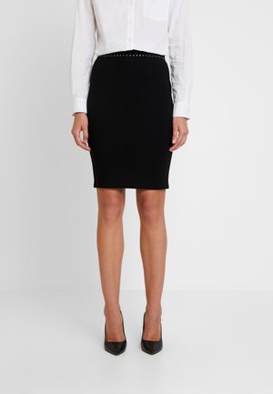 Femme portant une chemise blanche à manches longues, une jupe crayon noire cloutée et des talons aiguilles noirs à bout pointu, debout sur un sol blanc contre un mur blanc.