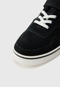 Sorte semska sko med hvite snører, med rund tå og en hvit midtsåle som er aksentuert med en tynn svart stripe. Teksturert overflate synlig.