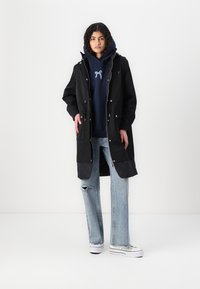 Svart parkajakke med hette, trykknapper og kontrasterende quiltet underpanel. Brukt over en marineblå hettegenser og lyseblå ripped jeans.